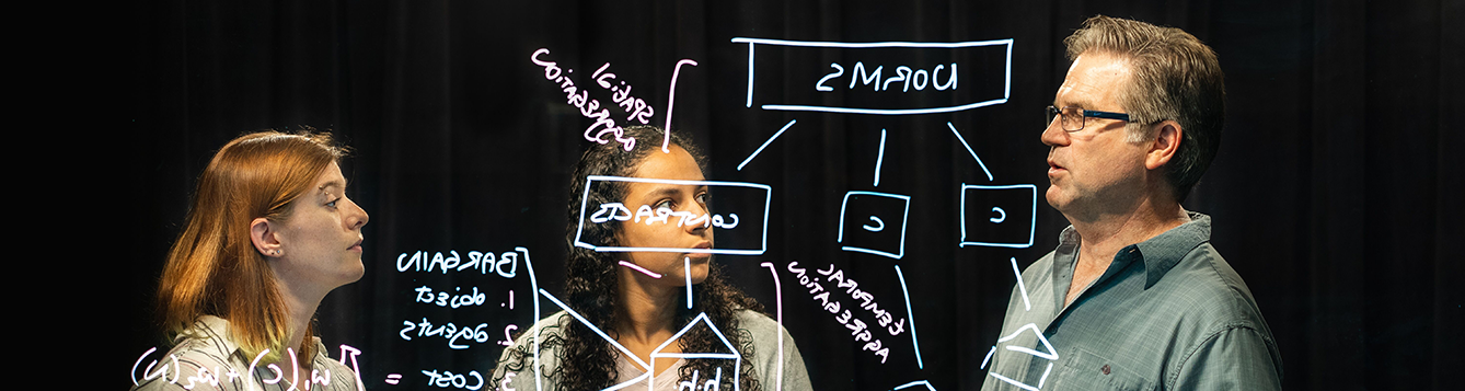 agricultural and biological engineering faculty member teaches two students science problems on a light board.