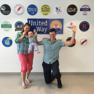 Two adults and a baby stand in front of a wall with many decorations, including a "United Way" sign.