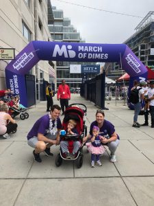 Two adults stand with a child in a stroller and a child standing on the ground, wearing purple shirts, outside of a March of Dimes purple arch way finish line.