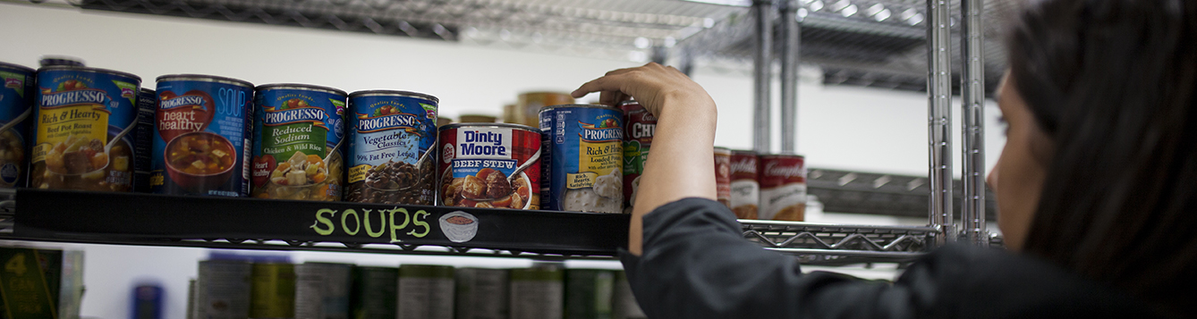 a volunteer reaches to stock cans of soup on a shelf at a food pantry