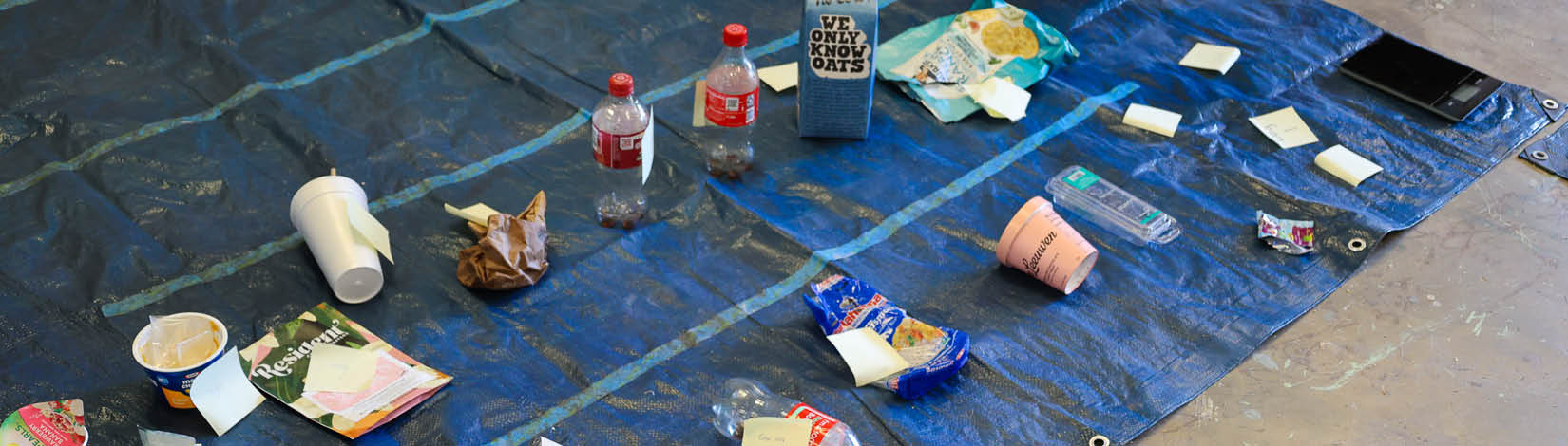 A variety of half empty food packages are arrayed on a blue tarp, tagged for cataloging. A small scale is on the side, used to weigh the objects to determine food waste amounts.