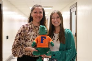 Two female graduate students smiling and holding the AEC AttaGator stuffed alligator