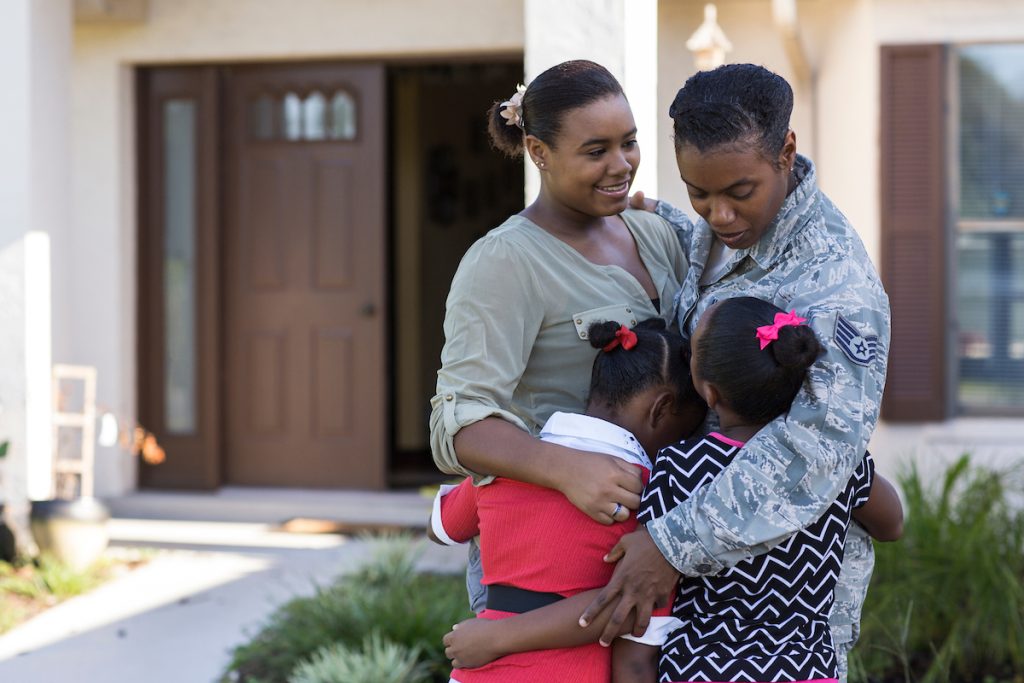 Active duty military mother hugs her three daughters in front of their home.