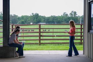 girl standing across from two peers giving her reasons. In the background is a paneled fence and cows.