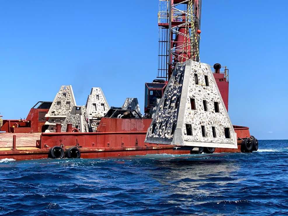Large concrete artificial reef structure in the foreground suspended from a crane by a yellow and black rope is slowing being lowered in the dark waters of the Gulf of Mexico.