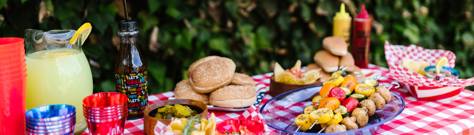 There is an image of a picnic table with red and white checkerboard table cloth. On the tablecloth from left to right there is an image of a pitcher of lemonade, red blue and red stacks of cups, a drinking bottle that says lets grill, a bowl of pickles, a plate of hamburger buns, a platter of veggie kabobs, a bowl of salsa, a plate of hot dog buns, a squeeze container of both mustard and ketchup and a paper basket with paper and food in it.