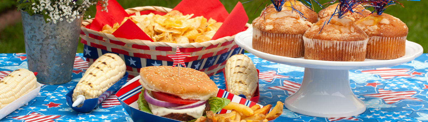 Image of chips, hamburger, corn and cake on a red white and blue table cloth