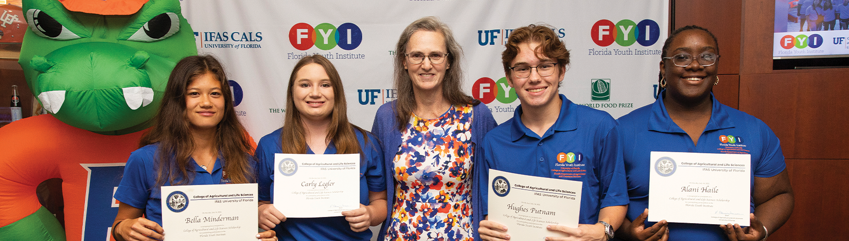 Four students, wearing blue polos and holding certificates, stand with the college dean in a floral patterned dress. Behind them, an inflatable alligator wearing a University of Florida shirt is placed next to a background with logos representing the UF/IFAS College of Agricultural and Life Sciences, Florida Youth Institute, and the World Food Prize Foundation.
