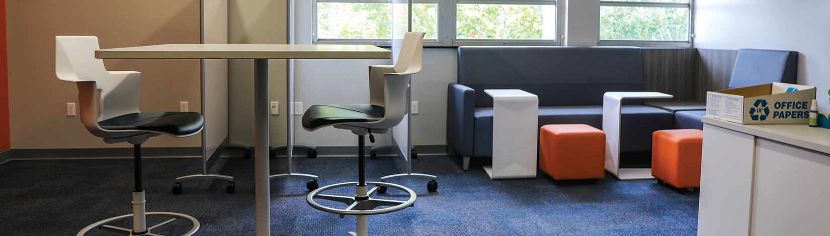 An empty study space with a high table and two chairs on the left, a gray couch on the right with small, white, portable desks.