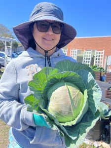 a woman standing outside in the sun holding a large cabbage.