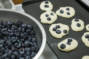 A bowl of blueberries and small blueberry pancakes cooking on a skillet. Photo taken 11-18-15