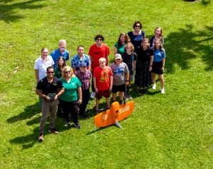 Students and presenters pose with a fixed wing drone while another drone takes their photo while in flight.