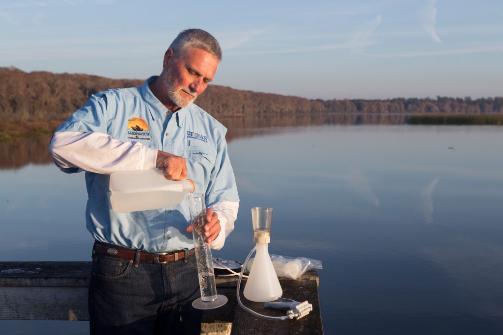 A man pours water from a jug into a cylindical tube as part of the water sampling process, with a lake as the backdrop 