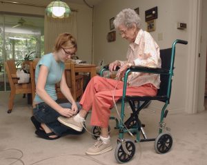 women helping an older woman in a wheelchair tie her shoes.