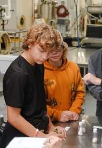 Two PK Yonge students--one wearing a black T-shirt and one wearing an orange hoodie sweatshirt--write on paper resting on a metal lab table | Food Science and Human Nutrition UF/IFAS
