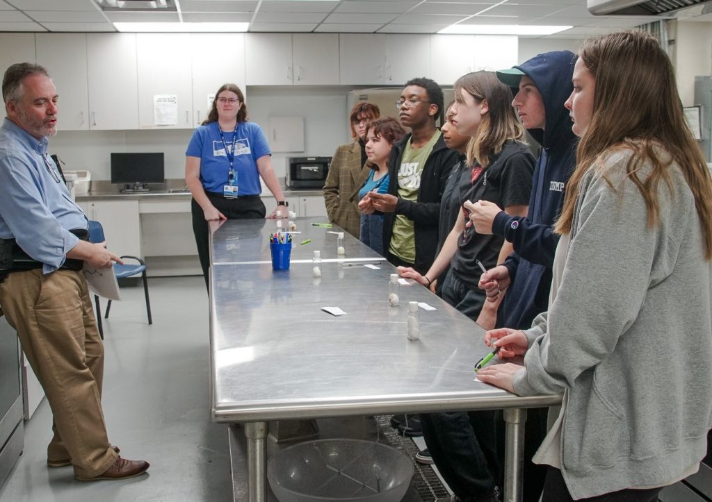 Professor in light blue collared shirt speaks to large group of high school students across a metal lab table, PK Yonge | Food Science and Human Nutrition UF/IFAS