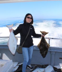 Dr. Liu wearing a dark top and holding a halibut in each hand while standing on a boat.