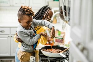 Female caregiver helping child pour tomato sauce into pan during meal preparation.