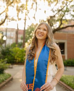 Julia wearing a blue graduation sash standing in front of a brown building and a tree.
