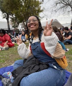 Rachel giving a peace sign while sitting on a blanket with people in the background.