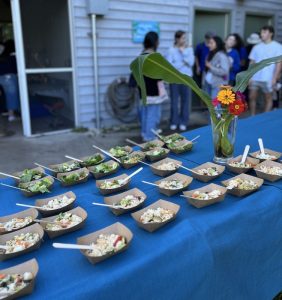 Trays of food lined up outside on a table with a blue tablecloth.