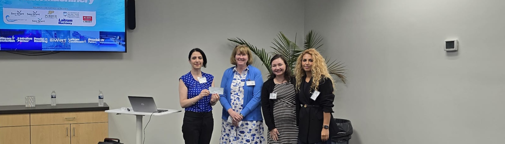 Rose standing with three women inside a conference room holding a check.
