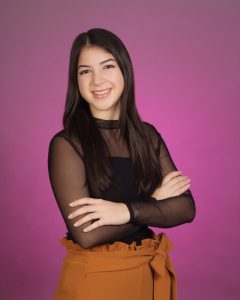 Maria Corredor wearing a black top and brown pants in front of a purple-pink background.