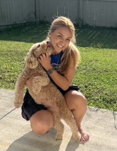 Dr. Farzad holding her light brown dog on a sidewalk with grass in the background.