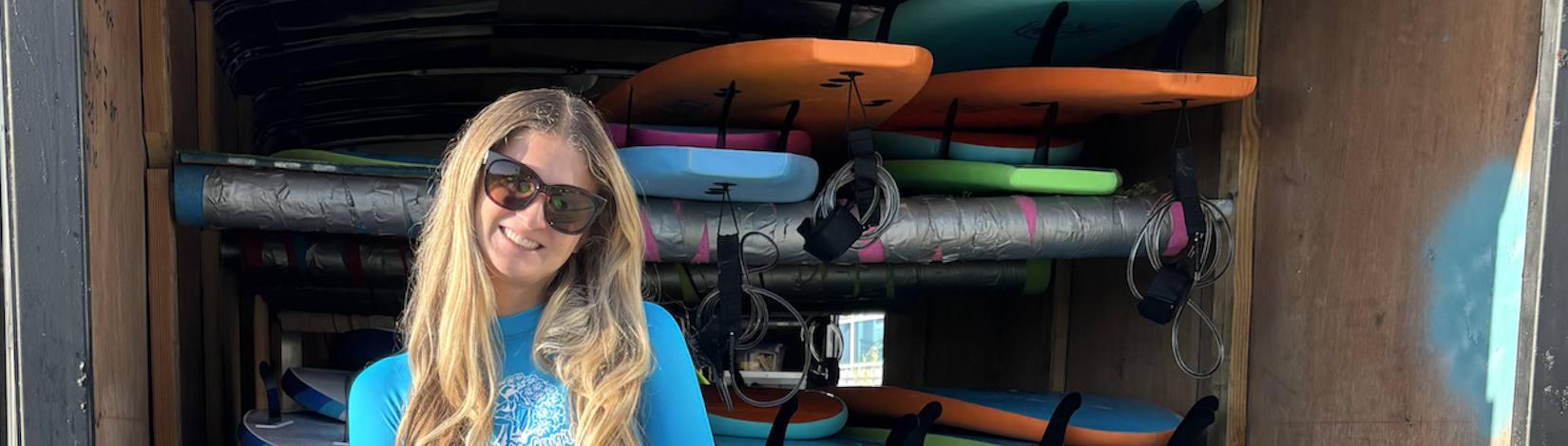 Avery coaching at a surf camp, wearing sunglasses in front of a truck full of surfboards.