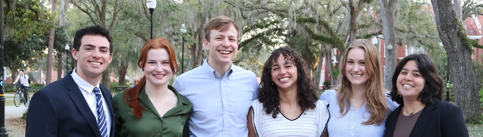 Six friends standing in a line in front of trees.