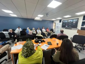  A professional meeting in progress with in-person attendees facing a large TV screen showing a video conference. There are banners on the wall, and the room has a U-shaped table arrangement.