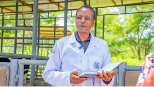 A livestock researcher wearing a lab coat stands in an open-air cattle facility, holding a notebook while speaking