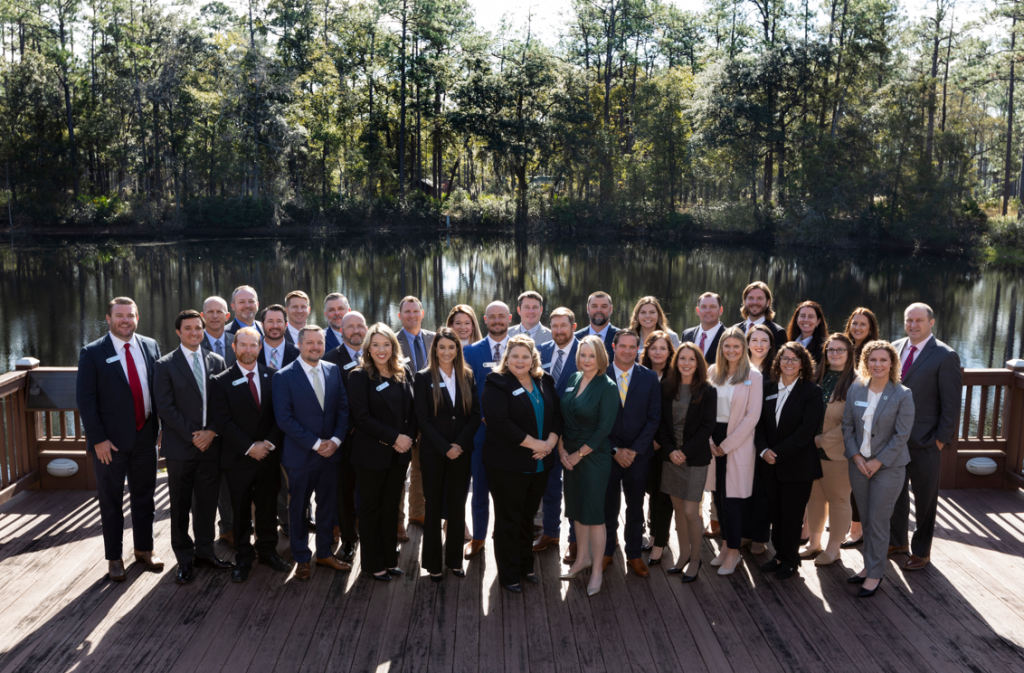 A group of 34 men and women in business attire pose together on a sunny day, standing on a wooden deck. There is a lake with trees in the background.