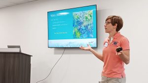 Lara standing up and presenting in front of TV screen with PowerPoint on it talking about tidal creeks