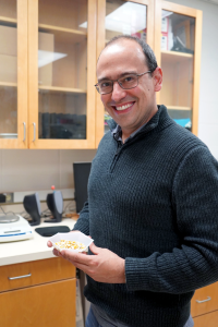 Dr. Marcio Resende stands in his lab holding maize kernels, wearing glasses and a navy sweater.