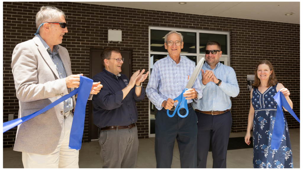 A ribbon-cutting ceremony outside a brick building. Five people stand in a row smiling—one man in the center holds oversized scissors cutting a wide blue ribbon while others clap and hold the ribbon.