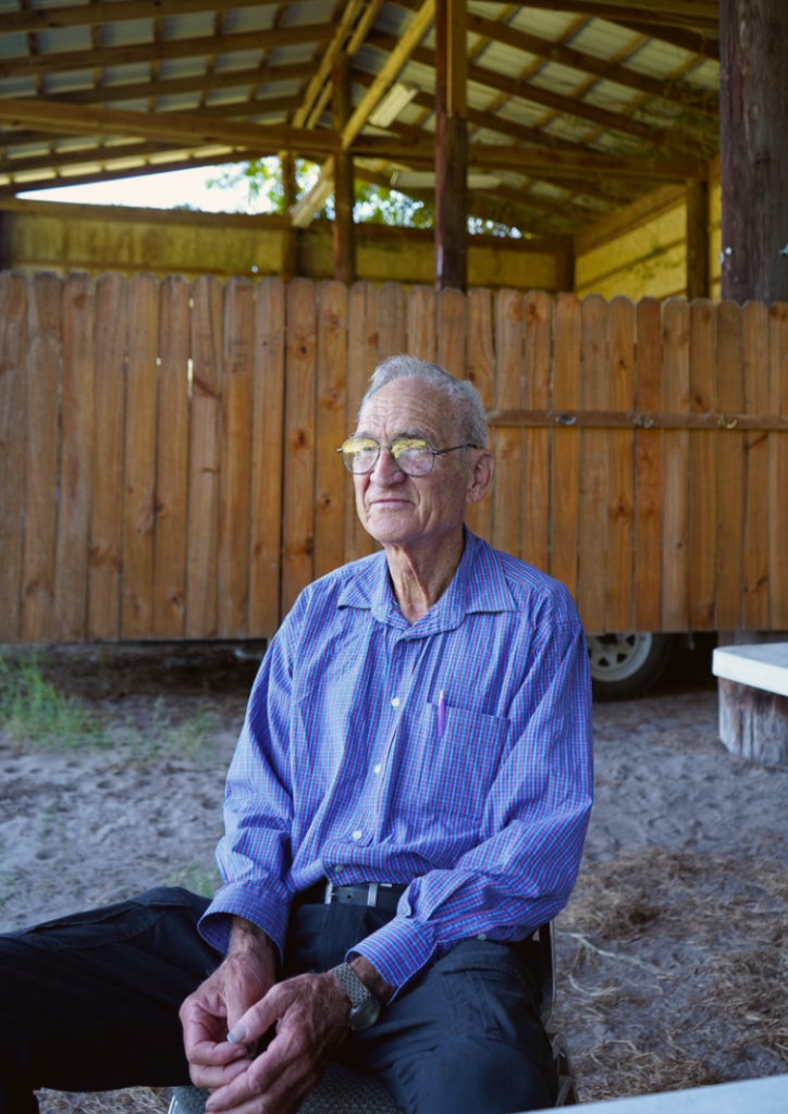 Dr. Paul M. Lyrene, wearing glasses and a blue checkered shirt, sits in front of a wooden fence under an open barn structure. His hands rest on his lap as he looks slightly off to the side with a calm, reflective expression.
