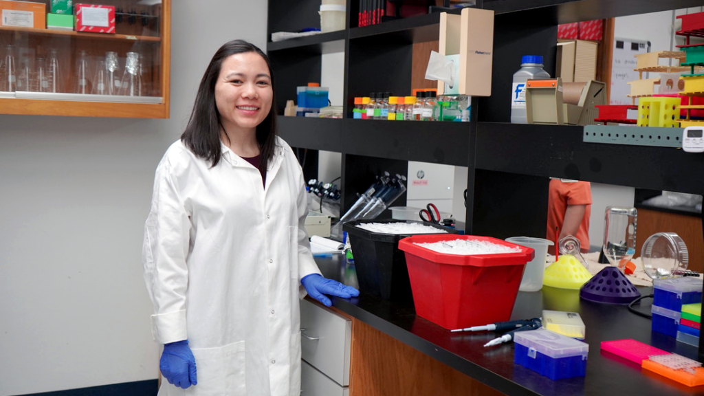 Woman in lab coat stands in front of counter smiling
