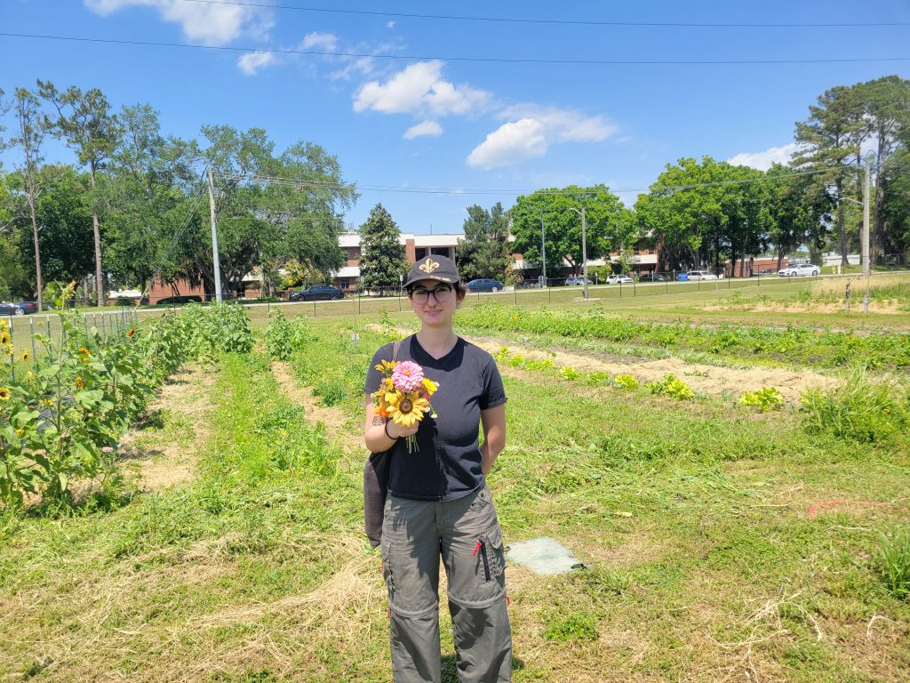 Alice Pekarsky stands in a black t-shirt and black jeans with a baseball cap and a freshly picked bouquet of wildflowers at the UF/IFAS Horticultural Sciences Teaching Farm