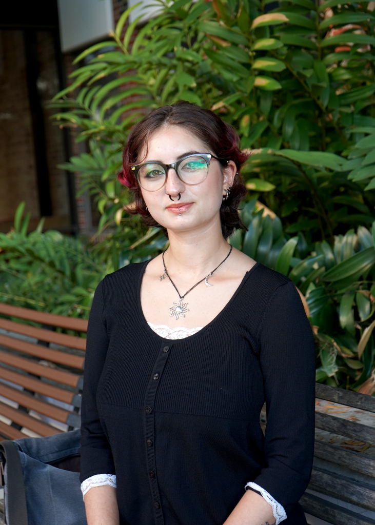 Young woman with brown-red hair in glasses sits on wooden bench with greenery in the background