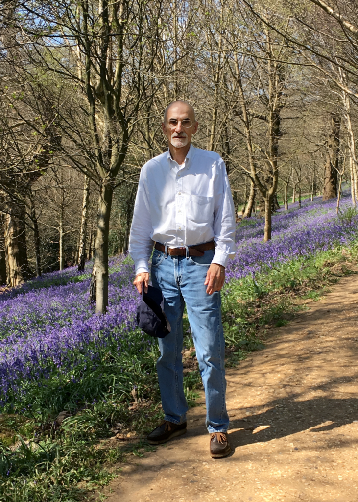 Andrew Hanson stands on a woodland path bordered by blooming purple wildflowers, wearing a white shirt, blue jeans, and glasses, holding a navy cap in his hand.
