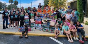 A large group of children and adults pose outdoors in front of a building, surrounded by tables and stacks of donated canned and packaged food. A central sign reads “Together WE CAN Make a Big DIFFERENCE” with a 4-H clover emblem. Participants wear 4-H-themed shirts and festive attire, including a cartoon turkey decoration, highlighting a Thanksgiving-themed community service event.