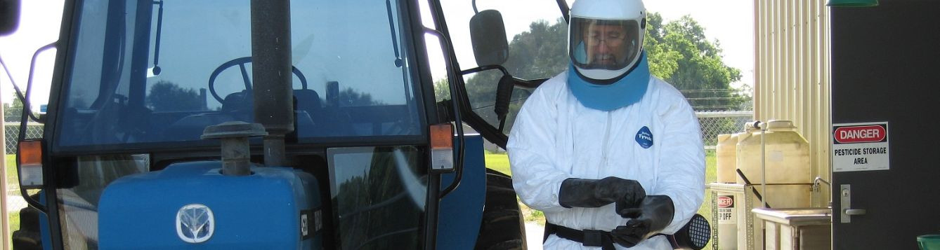 Properly suited pesticide applicator beside tractor sprayer ready to apply a chemical with danger signal word.