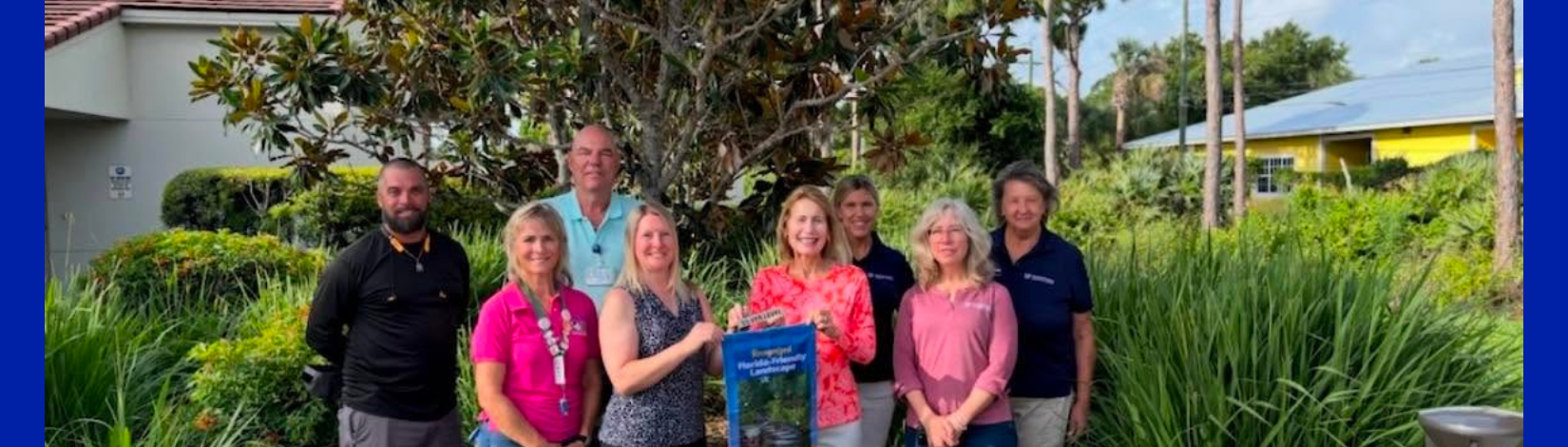 Six people are standing together in front of a Florida Friendly Landscape holding a flag that certifies the landscape as Florida Friendly.