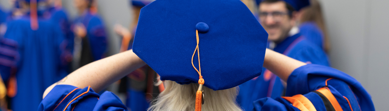 A graduate adjusts their blue cap adorned with a yellow tassel, surrounded by other graduates in similar regalia.