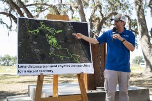 In an outdoor setting with trees, Hace Ellington stands next to an easel and points at a map