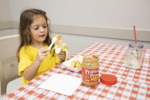 A little girl spreading peanut butter on a banana, sitting at a table with a red and white gingham pattern.