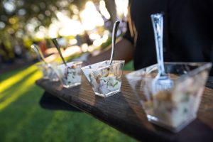 A server holding small containers of food with spoons in an outdoor setting.