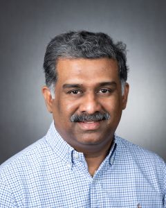 Doctor Paret wearing a light blue checkered shirt against a dark, softly lit background.