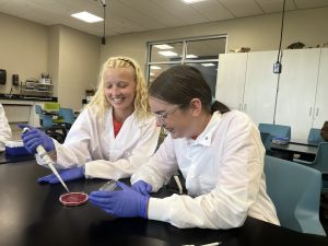 Two female students wearing lab coats and blue gloves working at a lab table with a petri dish and a pipette.